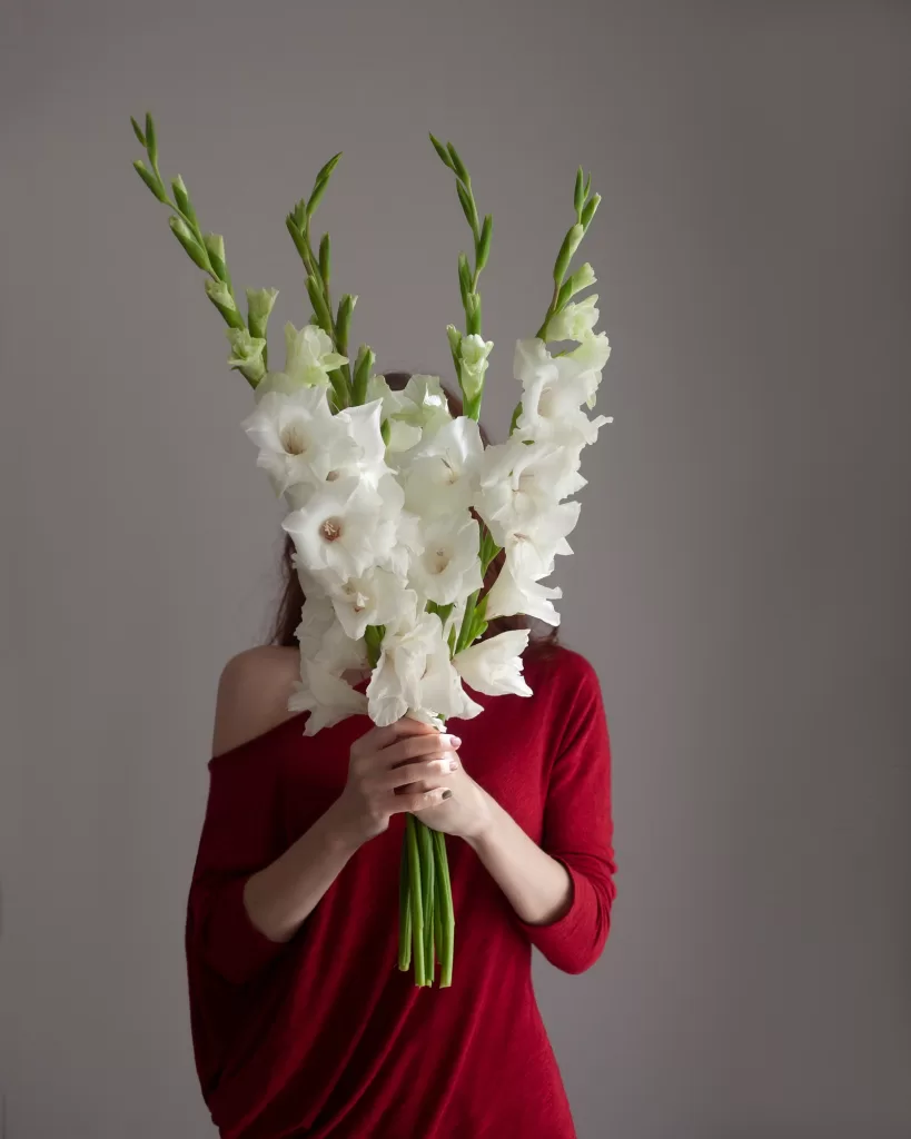 a red-haired girl in a red sweater holds a bouquet of white gladioli covering her face with them